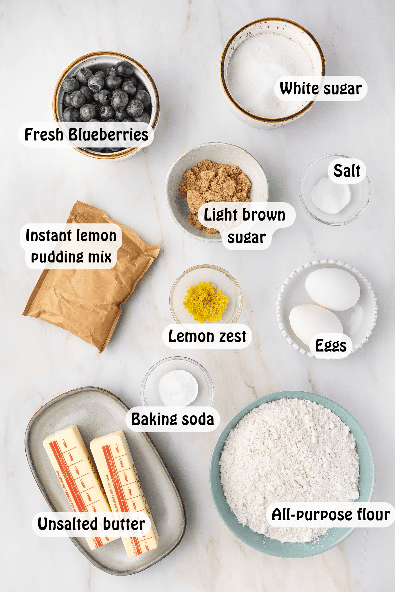 Ingredients for lemon blueberry cookies, including unsalted butter, white sugar, light brown sugar, instant lemon pudding mix, eggs, all-purpose flour, baking soda, salt, fresh blueberries, and grated lemon zest, neatly placed in a bowl.