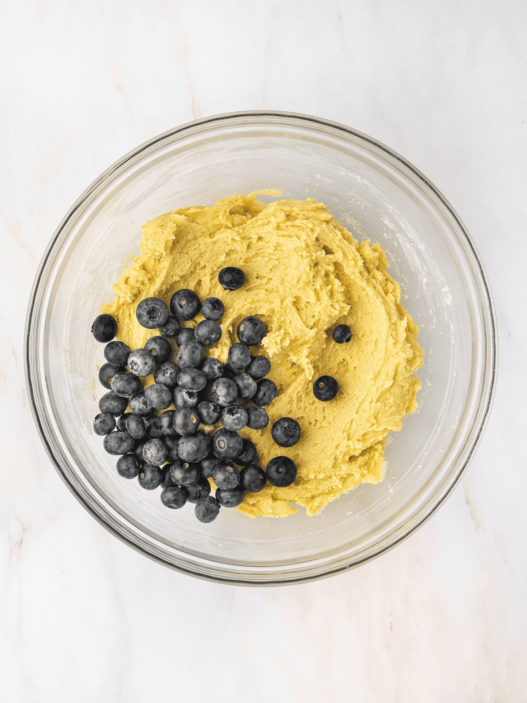 Fresh blueberries and grated lemon zest being gently folded into cookie dough in a mixing bowl.