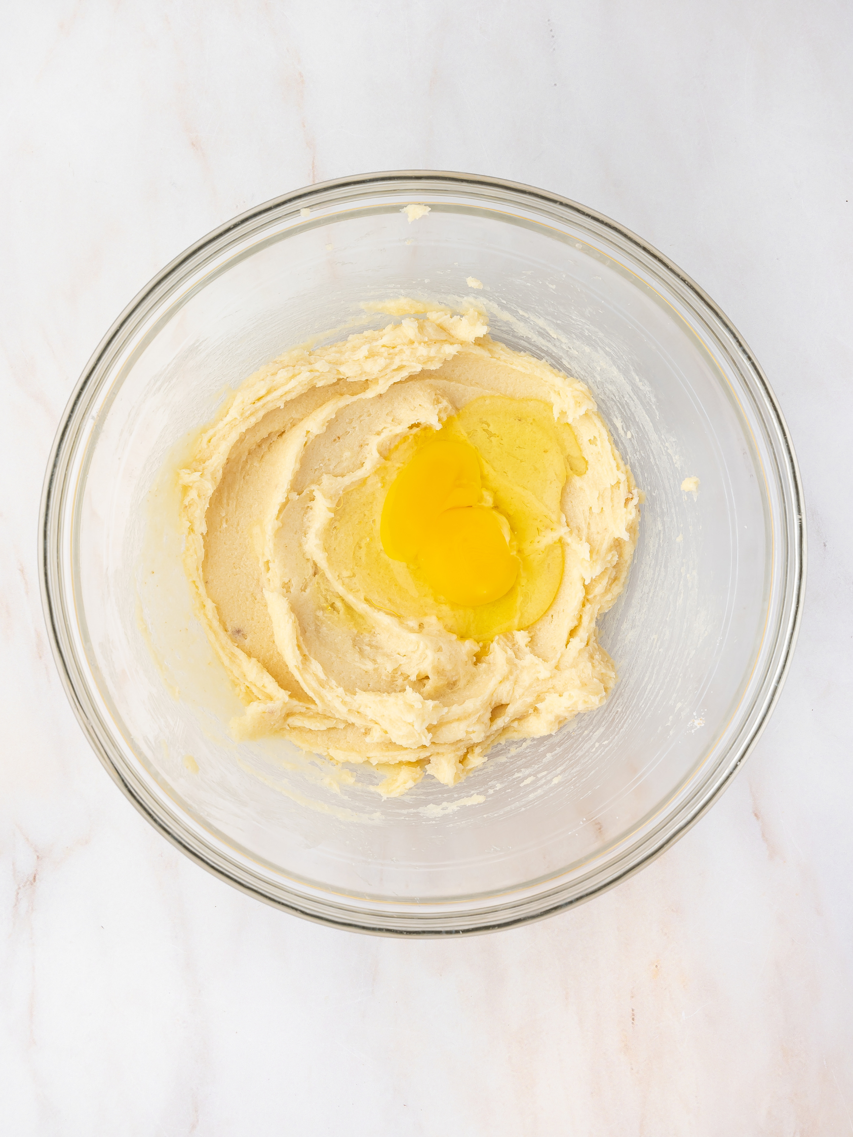 Creamed butter and sugar mixture in a glass bowl with an egg being added one at a time, ready to be mixed into the dough.