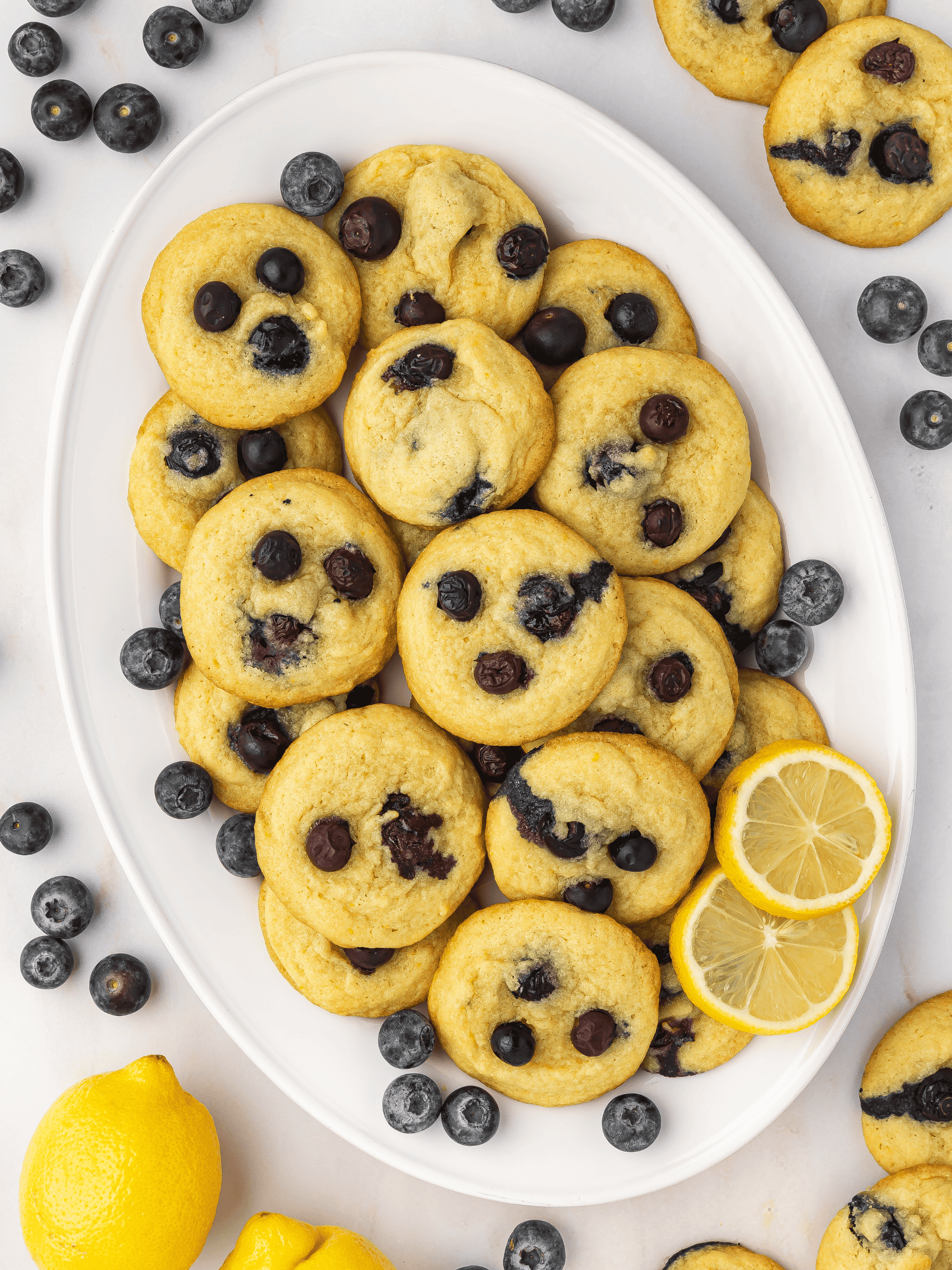 Lemon blueberry cookies arranged on a white ceramic platter, with fresh lemon slices and blueberries scattered around for garnish