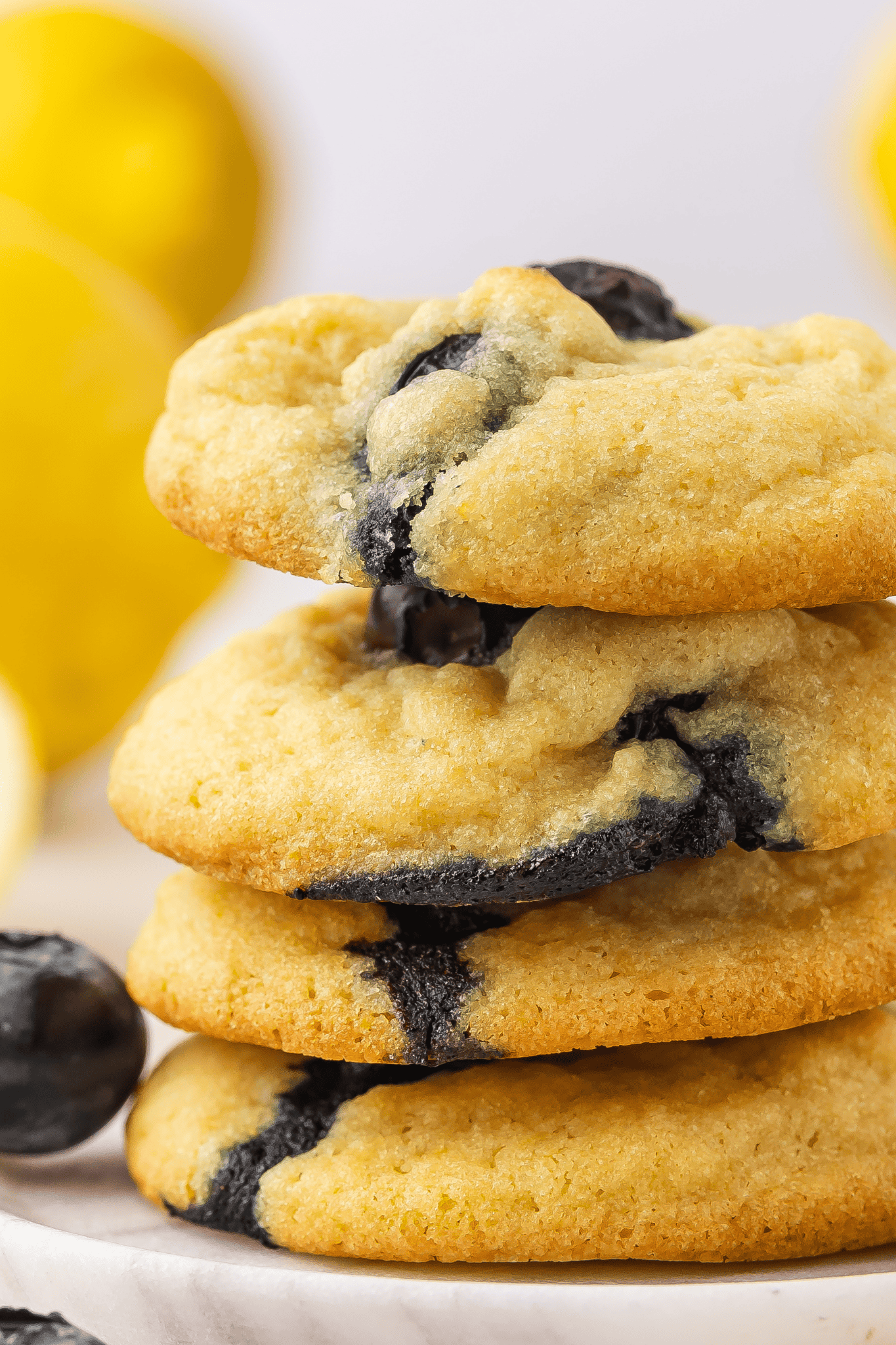 Four lemon blueberry cookies stacked one above the other, with fresh blueberries placed on the sides and a lemon in the background.