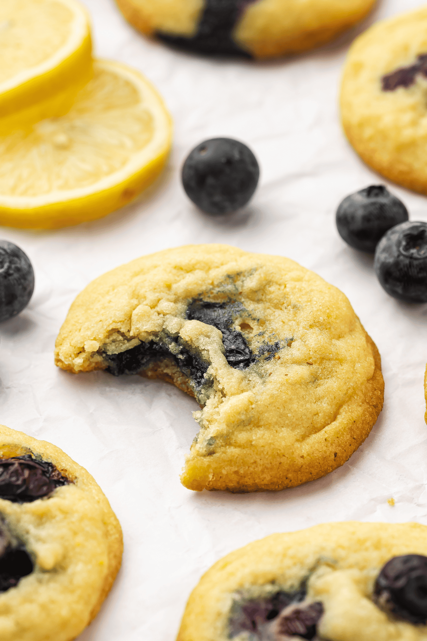 Lemon blueberry cookies with a bite taken from one, scattered around on a table with some blueberries at a 45-degree angle, showcasing their soft and chewy texture.