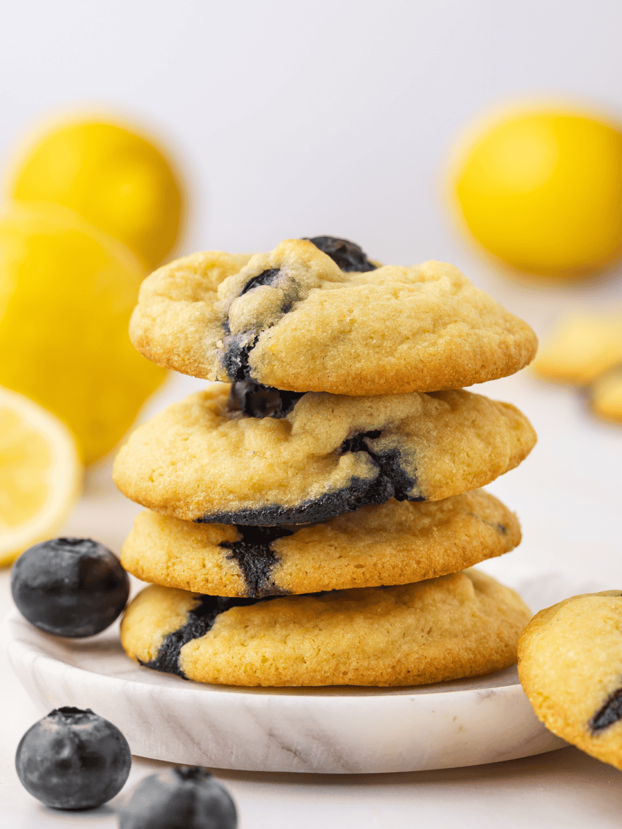 Four lemon blueberry cookies stacked one above the other, with fresh blueberries placed on the sides and a lemon in the background.