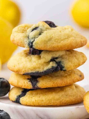 Four lemon blueberry cookies stacked one above the other, with fresh blueberries placed on the sides and a lemon in the background.