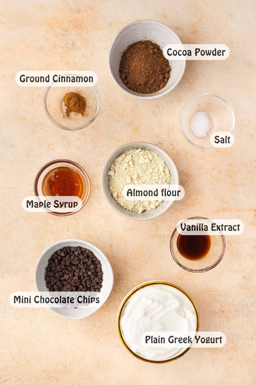 A flat lay of various ingredients in small bowls, including Greek yogurt, almond flour, cocoa powder, maple syrup, vanilla extract, cinnamon, and a pinch of salt, beautifully arranged on a clean surface.