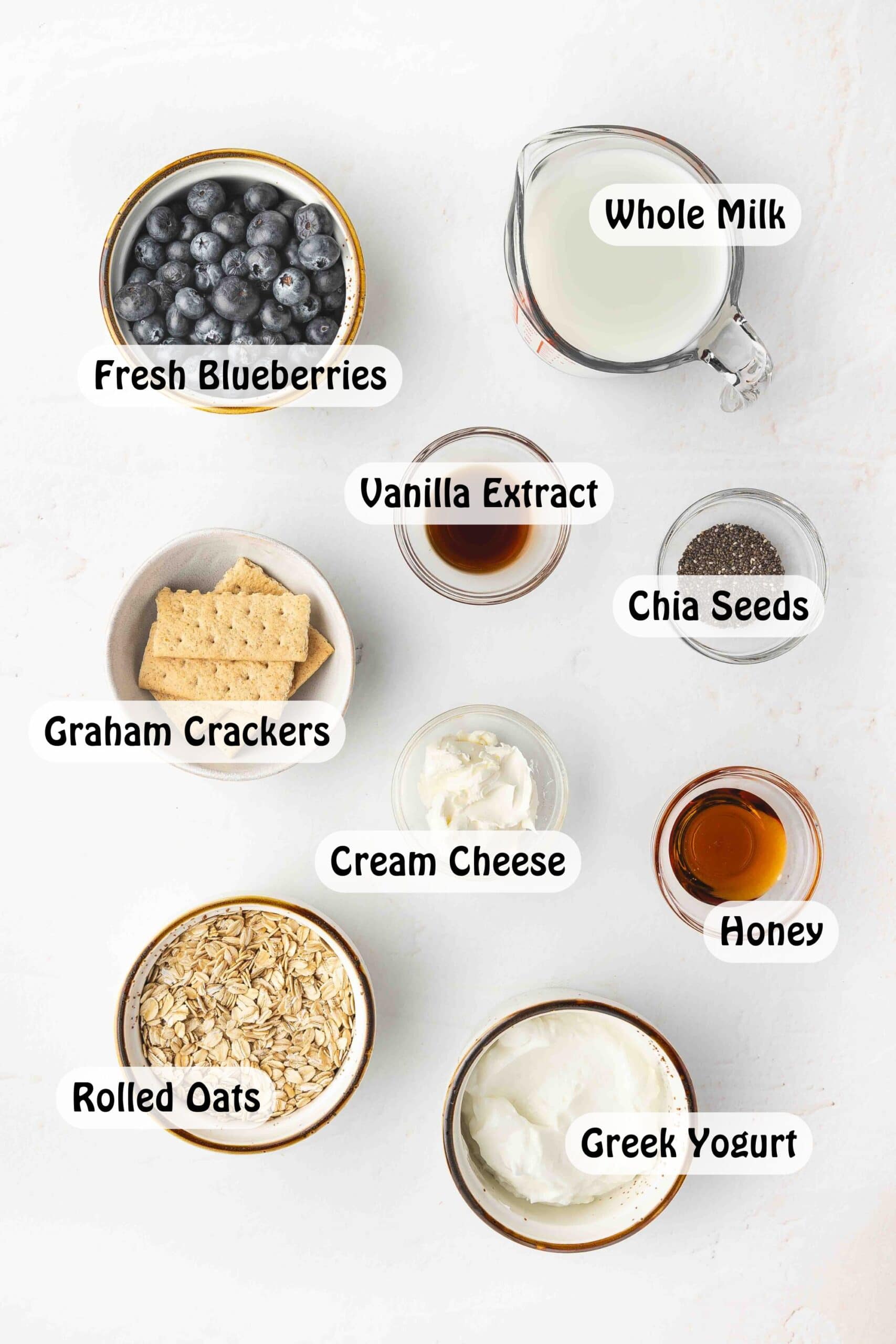 Ingredients for Blueberry Cheesecake Overnight Oats on marble background including rolled oats, chia seeds, blueberries, Greek yogurt, cream cheese, honey, and graham crackers.