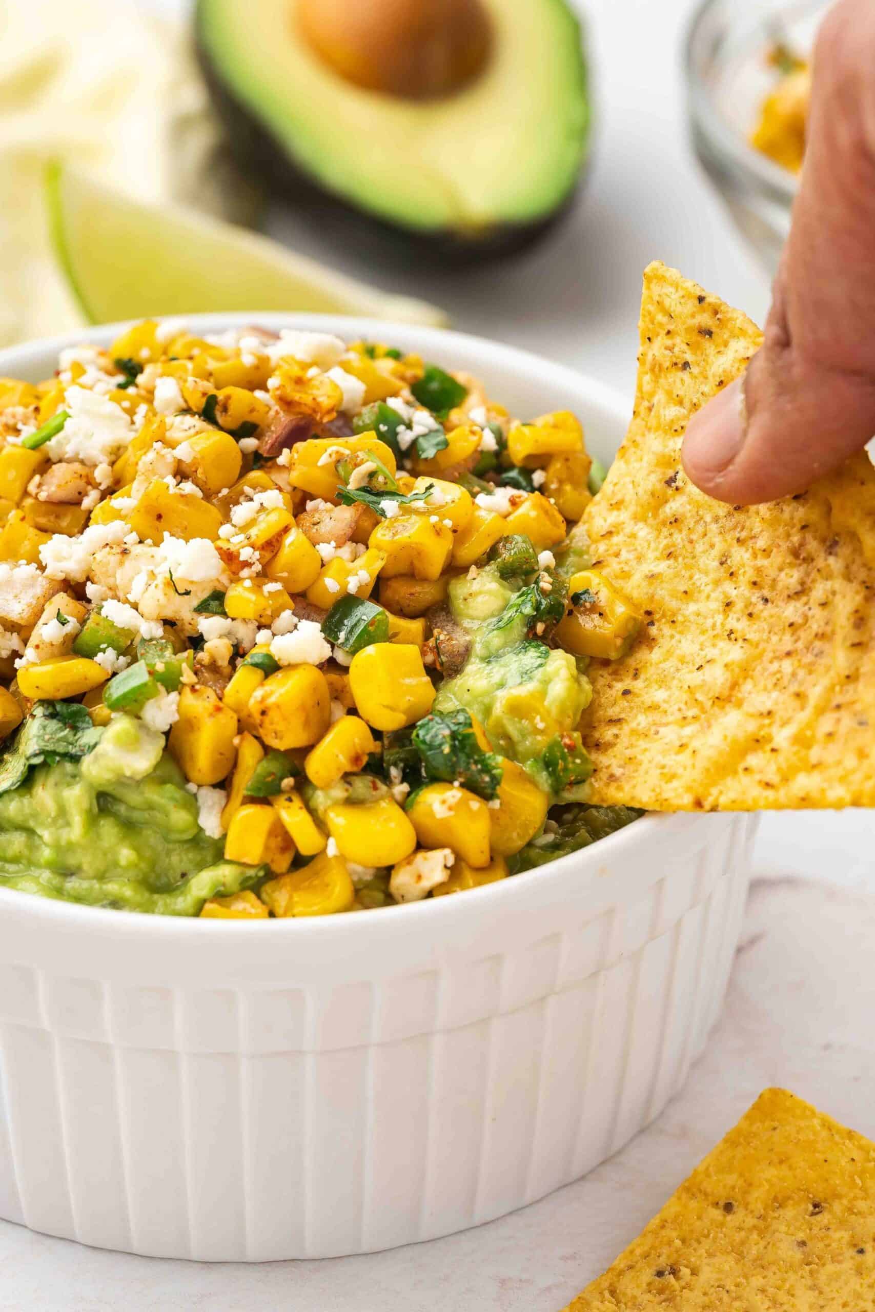 Tortilla chip scooped with Mexican street corn guacamole with a side of cilantro and lime wedges.