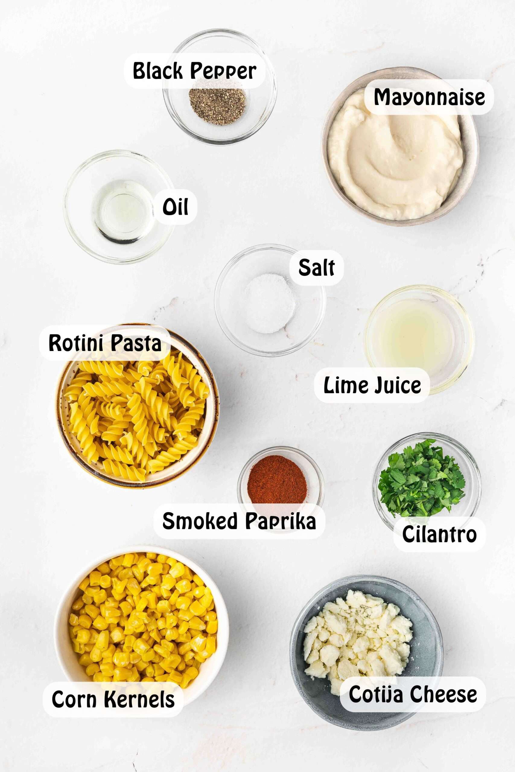 A top-down view of ingredients for Street Corn Pasta Salad, including rotini pasta, fresh corn kernels, mayonnaise, cotija cheese, lime juice, smoked paprika, salt, black pepper, and chopped cilantro.