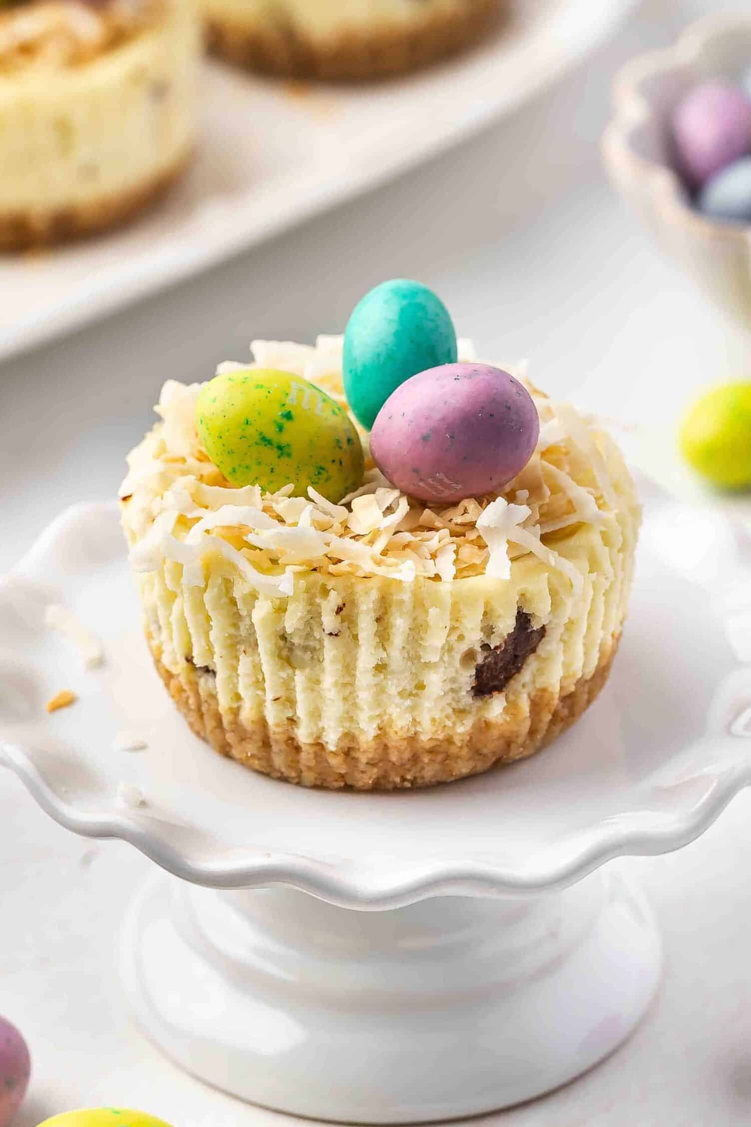 Mini Easter cheesecakes garnished with candy-coated eggs, displayed on a small cake stand.