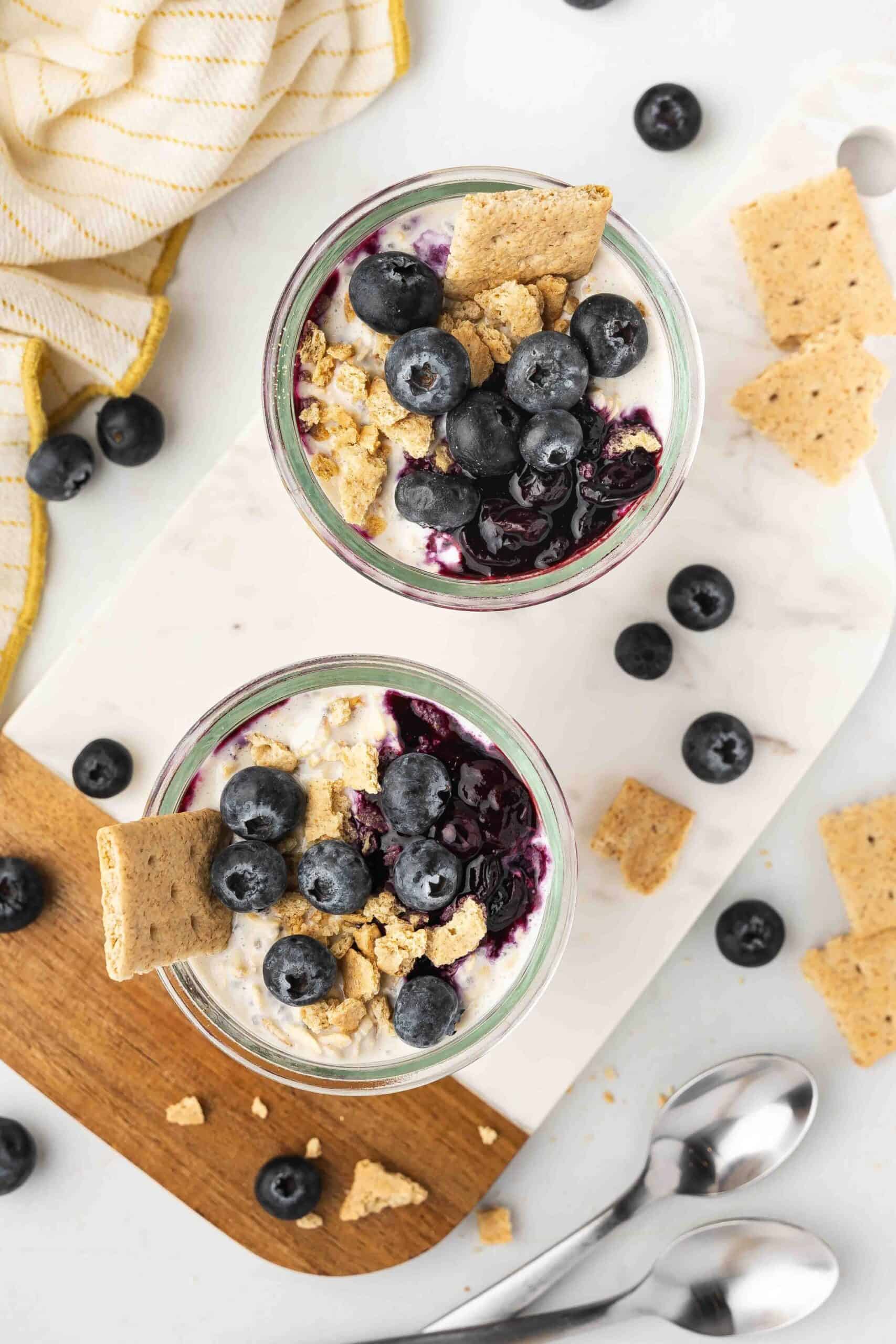 Top angle shot of two cups of Blueberry Cheesecake Overnight Oats with scattered blueberries.