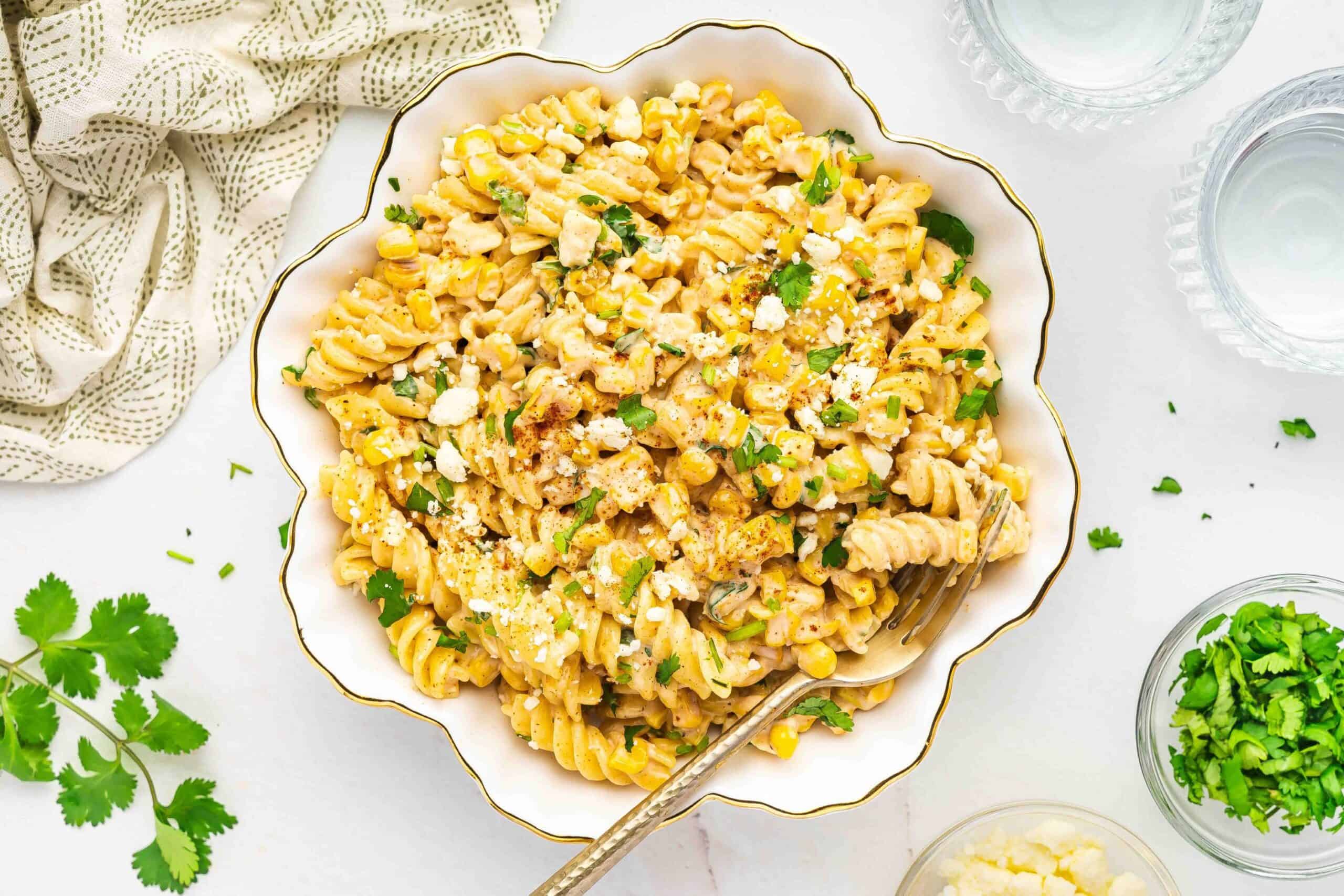 A bowl of street corn pasta salad surrounded by fresh ingredients like corn, lime, and cilantro.