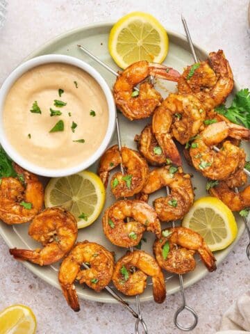 Oven-baked shrimp skewers with spicy garlic aioli and lemon slices on a plate.