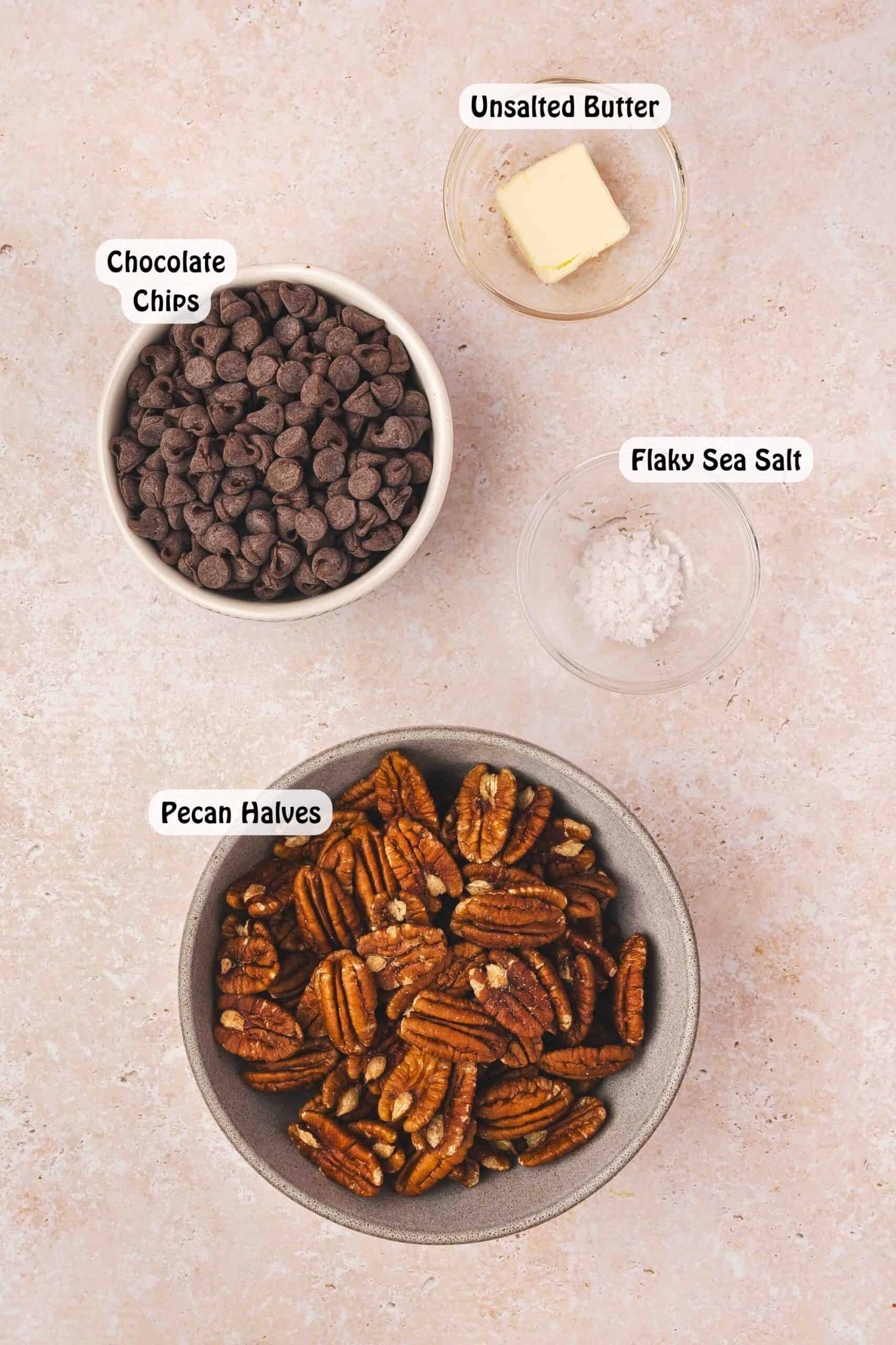 Ingredients for half-dipped chocolate-covered pecans: pecan halves, semi-sweet chocolate chips, unsalted butter, and flaky sea salt on a countertop.