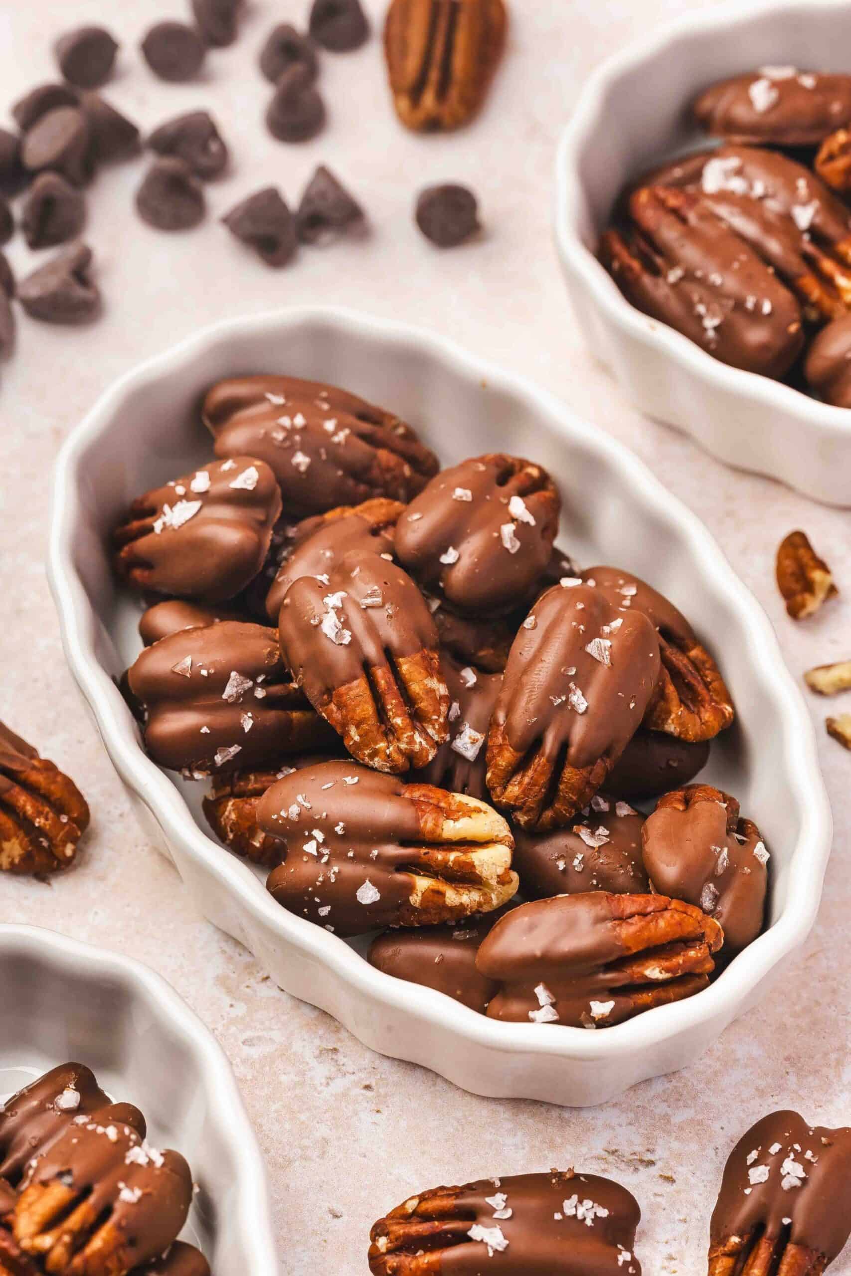 Half-dipped chocolate-covered pecans in a white ceramic bowl on a kitchen table.