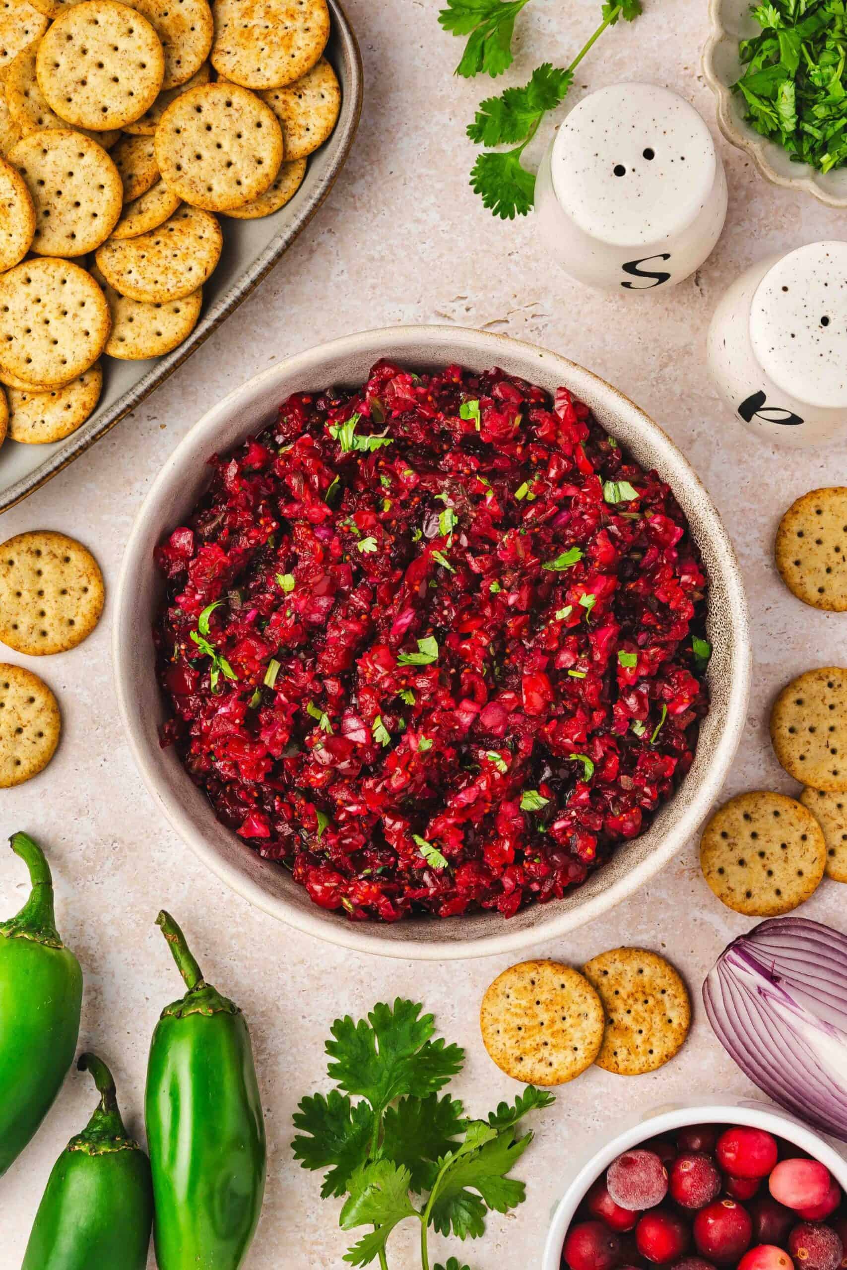 Colorful cranberry jalapeño salsa in white bowl with multigrain crackers on side