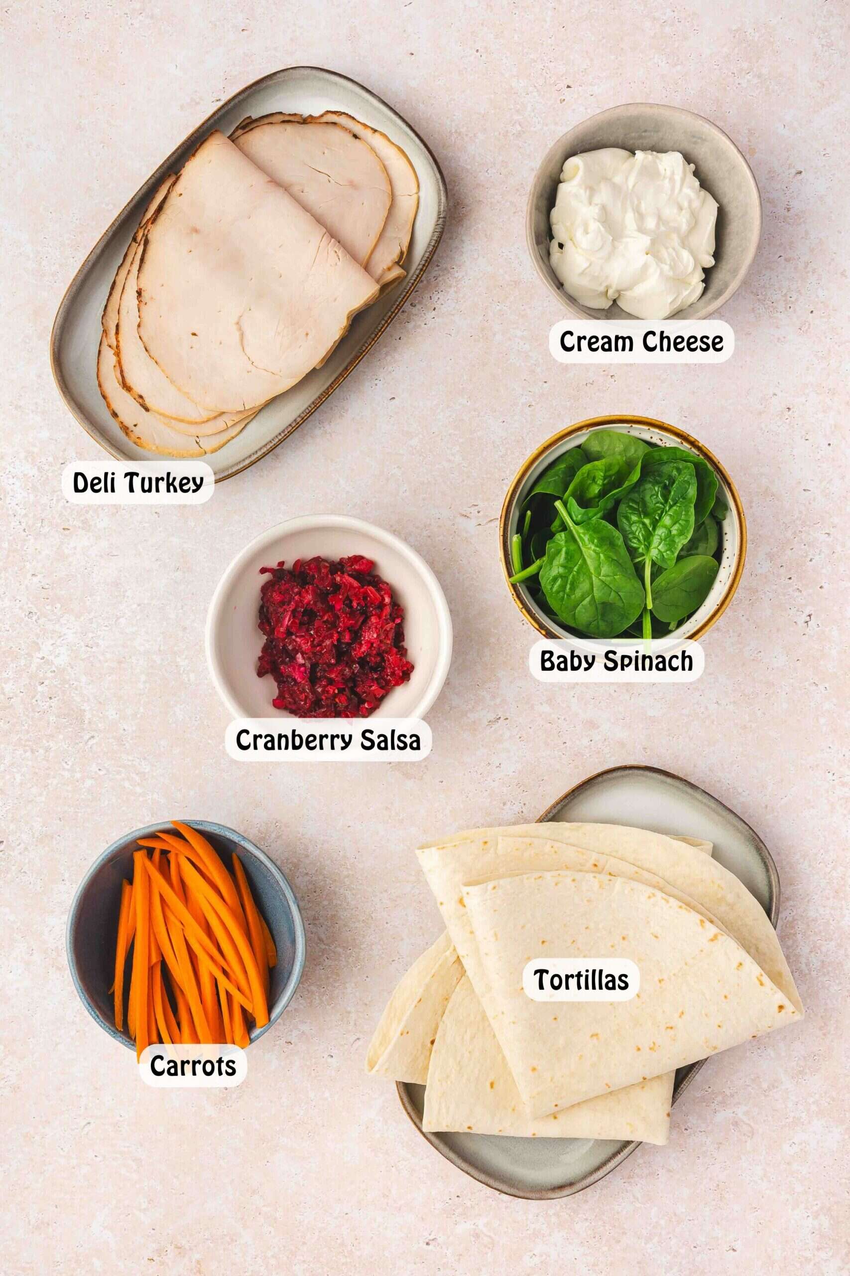 Ingredients for turkey roll-ups with cranberry cream cheese and fresh veggies on a cutting board, including tortillas, turkey slices, cream cheese, cranberry jalapeño salsa, spinach, and carrots.
