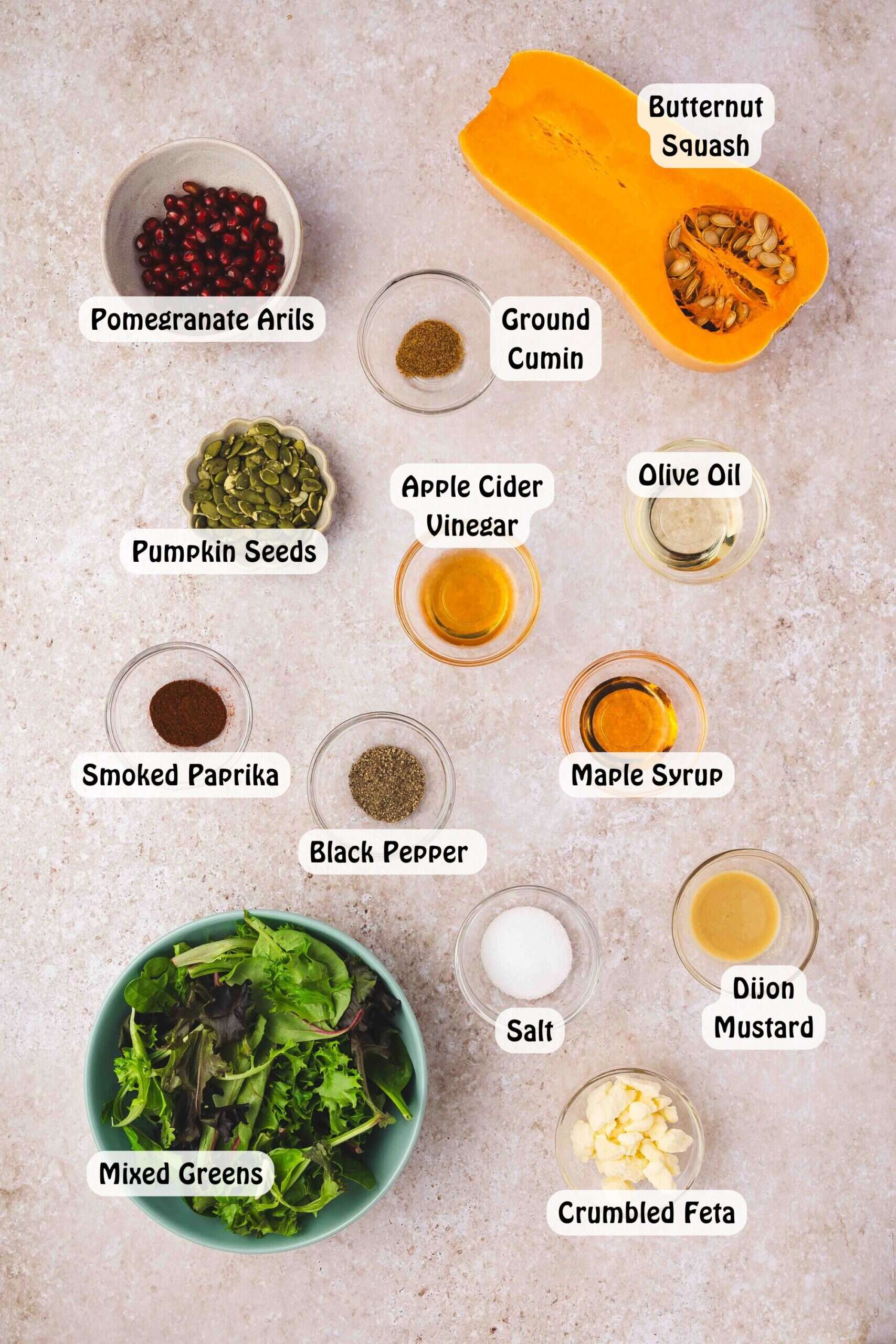 Ingredients for butternut squash feta salad laid out on a counter: ½ butternut squash, olive oil, smoked paprika, cumin, salt, pepper, mixed greens, pomegranate seeds, feta, pumpkin seeds, and salad dressing ingredients