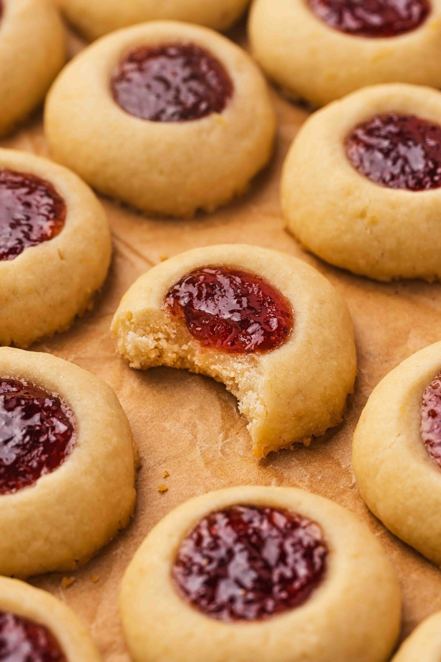 Rustic baking setup with raspberry thumbprint cookies scattered on parchment paper beside a small jar of jam.