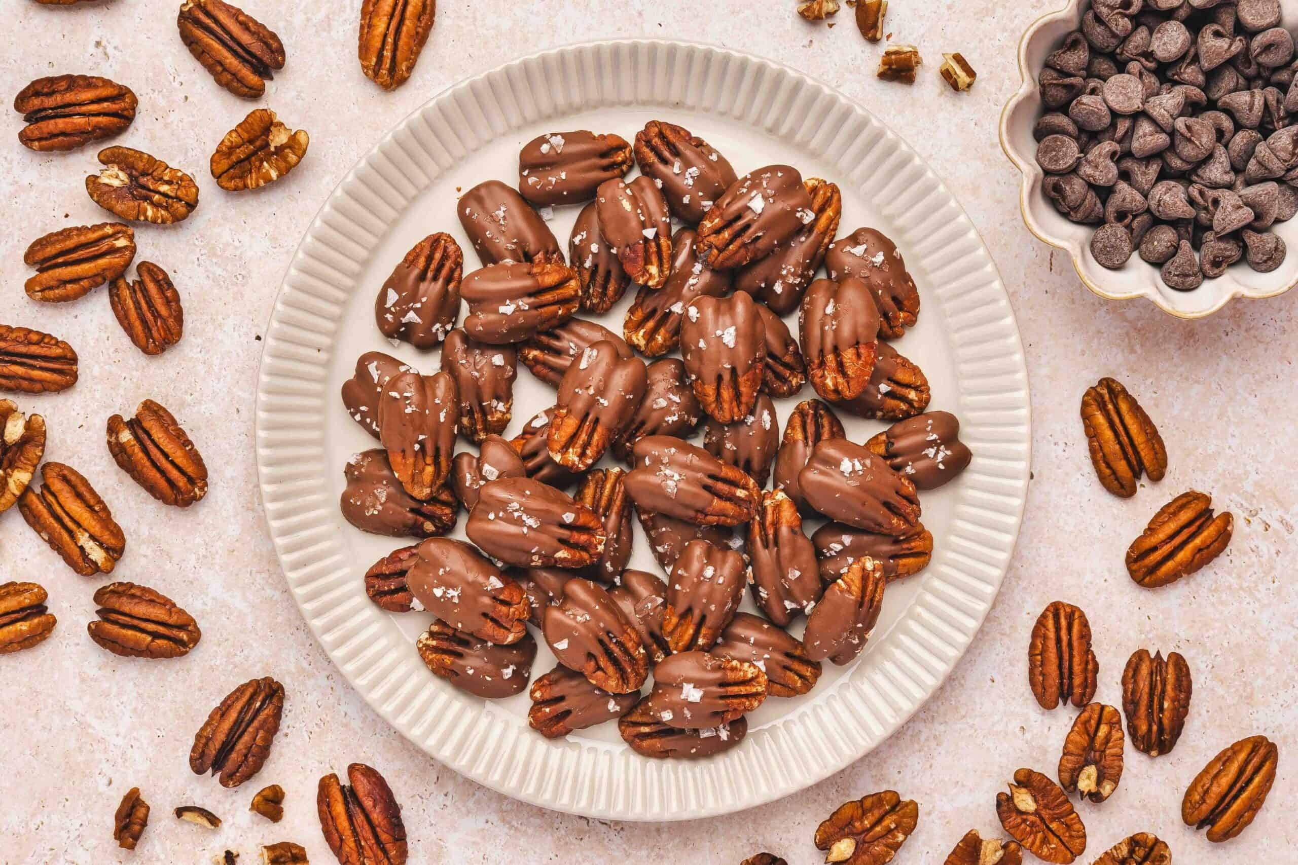 Half-dipped chocolate-covered pecans arranged on a white ceramic serving plate with flaky sea salt.
