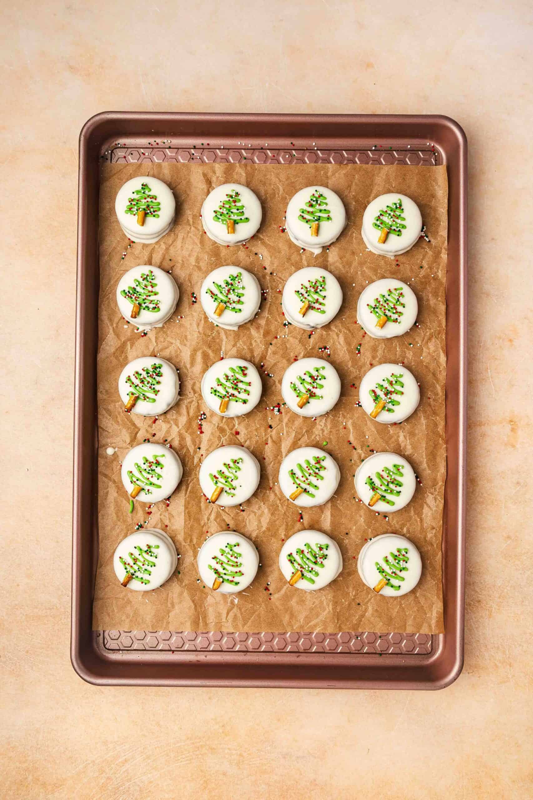 Refrigerating decorated white chocolate covered Oreos until green drizzle and pretzel trunks are set.