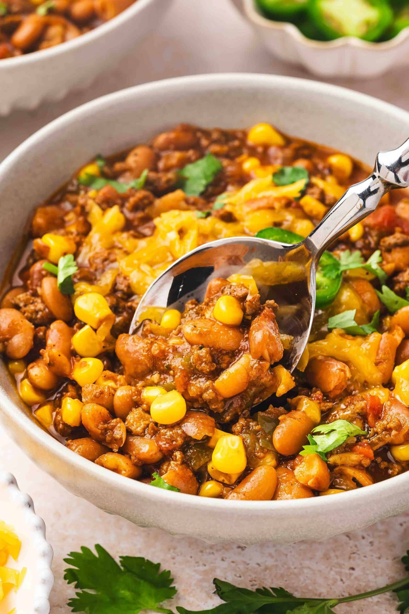 Close-up of spicy beef chili in a bowl with beans, corn, and fresh green toppings.